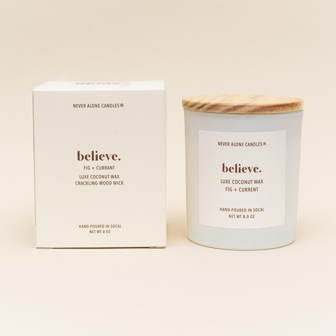 Hand-poured coconut wax candle with a crackling wood wick, scented with Fig and currant, next to a white box with the message 'Believe".