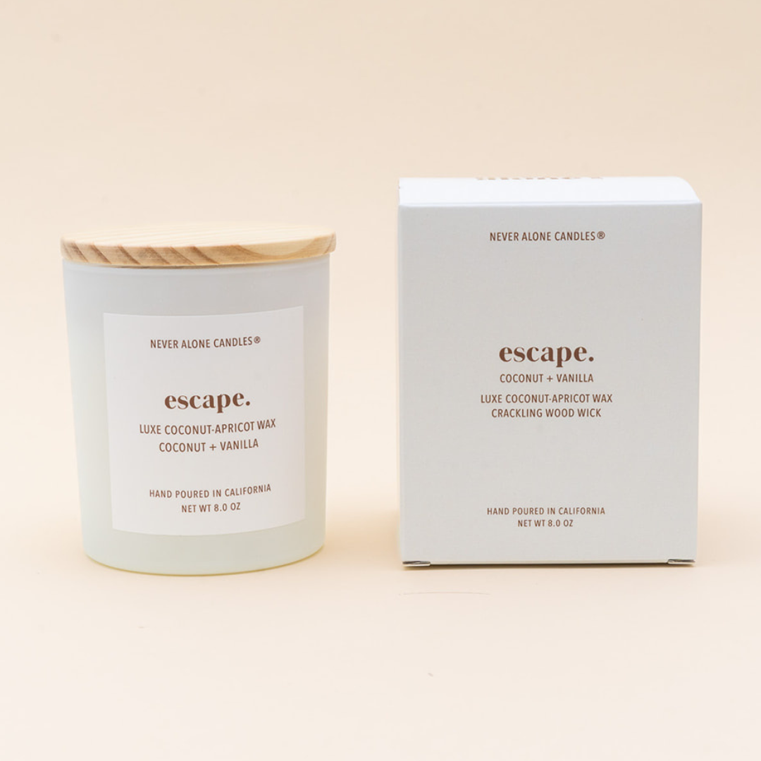 A candle jar with a light wood lid and a white label next to its matching white product box, both branded Never Alone Candles and labeled 'escape. Coconut + Vanilla'.