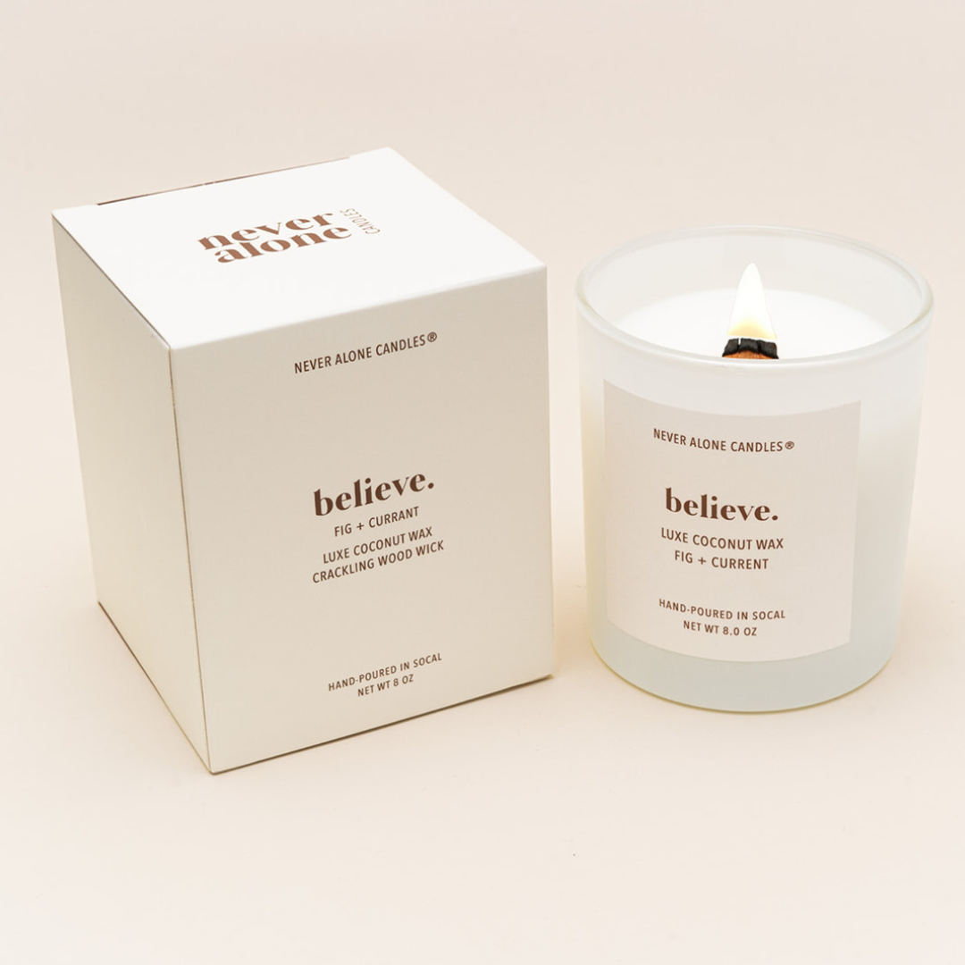Hand-poured coconut wax candle with a crackling wood wick, scented with fig and currant, next to a white box with the message 'believe'.