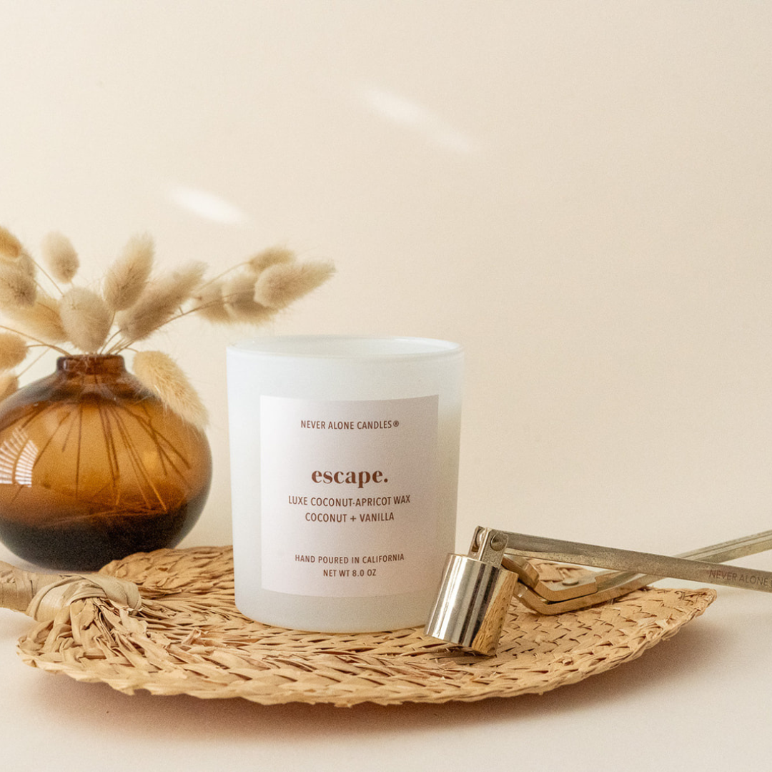 A matte white candle jar labeled 'escape' by Never Alone Candles, styled on a woven tray with a gold wax dipper and dried grass in an amber glass vase against a warm beige background.