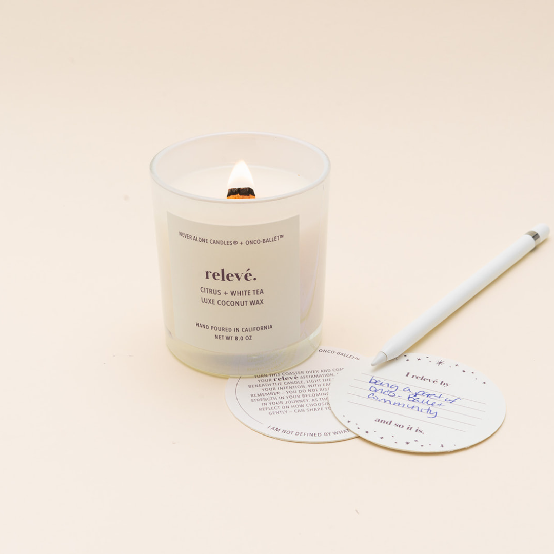 A lit white coconut wax candle in a clear glass jar with a white label reading 'relevé' and 'Citrus + White Tea'. A white pen and a manifestation card are placed beside it on a warm cream background.