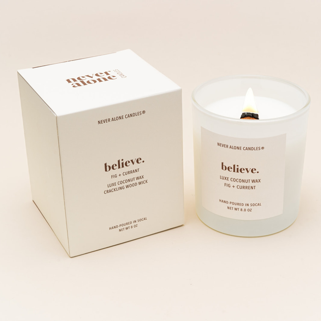 Hand-poured coconut wax candle with a crackling wood wick, scented with fig and currant, next to a white box with the message &#39;believe&#39;.